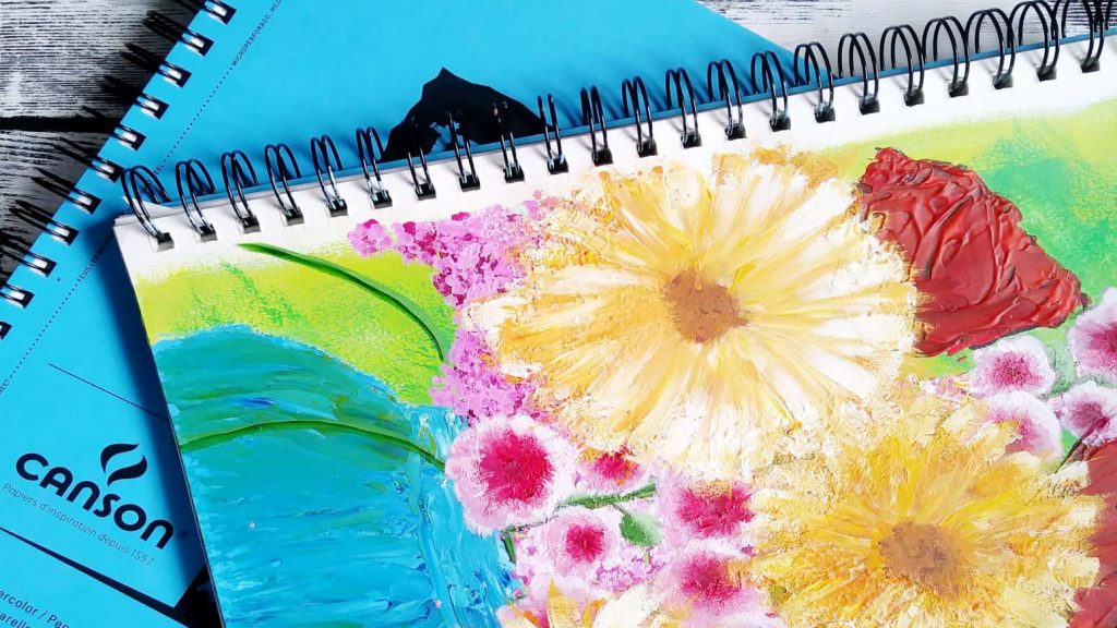 A Canson mixed media paper journal opened to a painting of a blue vase with assorted brightly colored flowers on a lime green background.