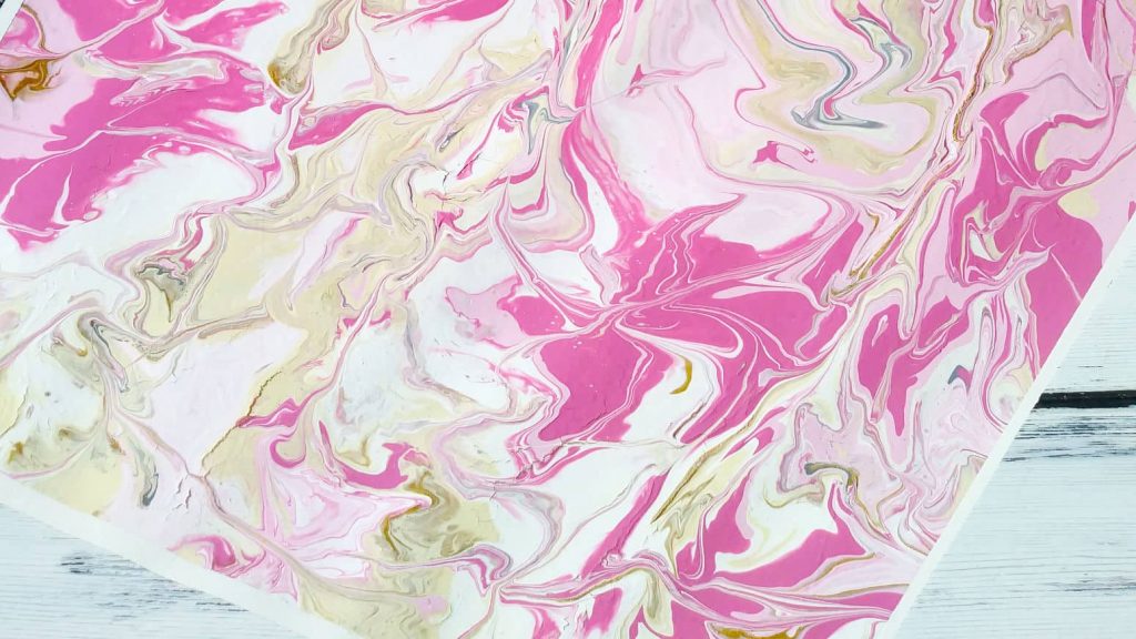 A pink, gold, white, and grey swirled painting on paper, with acrylics, by using the paint pour method.