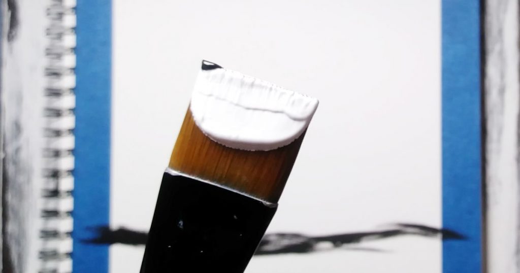 A flat brush with mostly white paint on it and a tiny bit of black paint on the tip of one corner