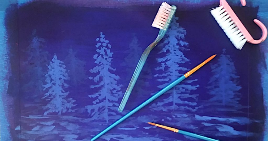 Dark blue landscape painting with distant snowy trees, snow covered ground, with two round paintbrushes, a toothbrush, and a nailbrush sitting on top which are the tools used to paint snowflakes.