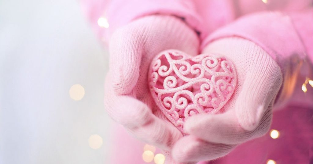 A pink scroll-work heart ornament held in cupped hands covered in pink gloves. A great reference photo to add to the list of possible Valentines paint night ideas!