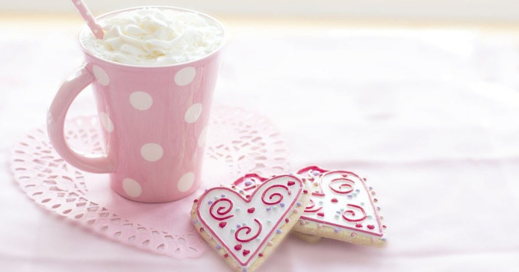 A pink and white polka-dot mug filled with whipped cream and a pink and white striped straw with two decorated heart-shaped cookies
