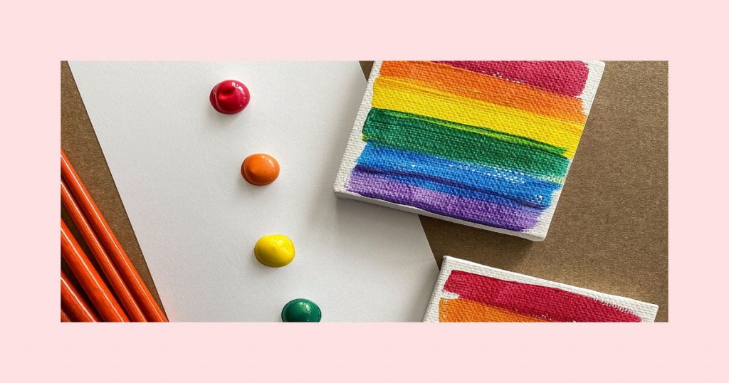 Two mini DIY rainbow art canvases covered in stripes of rainbow colored paint and a palette with blobs of rainbow colored paint on it