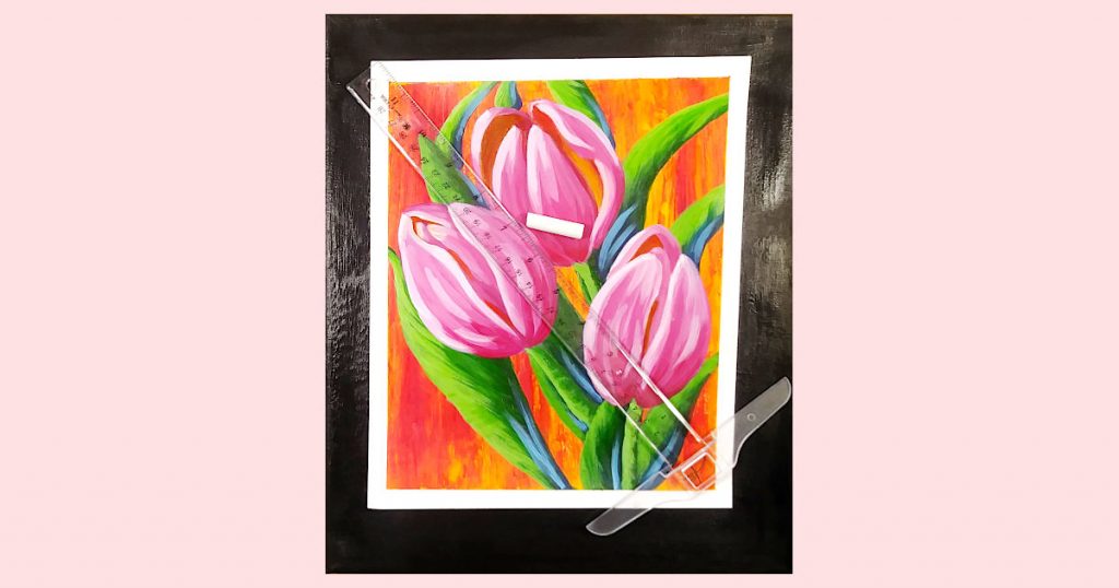 The pink tulip paper painting on top of the black painted canvas with a piece of white chalk and a ruler