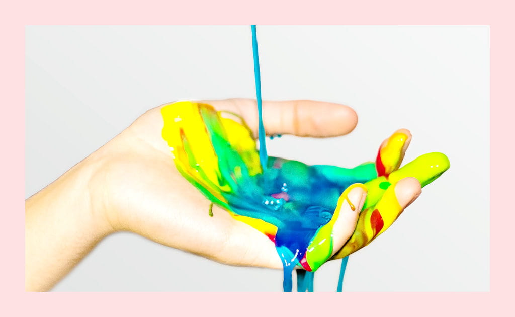 Bright yellow, blue, green, and red acrylic paint being poured onto an adult's upturned palm.