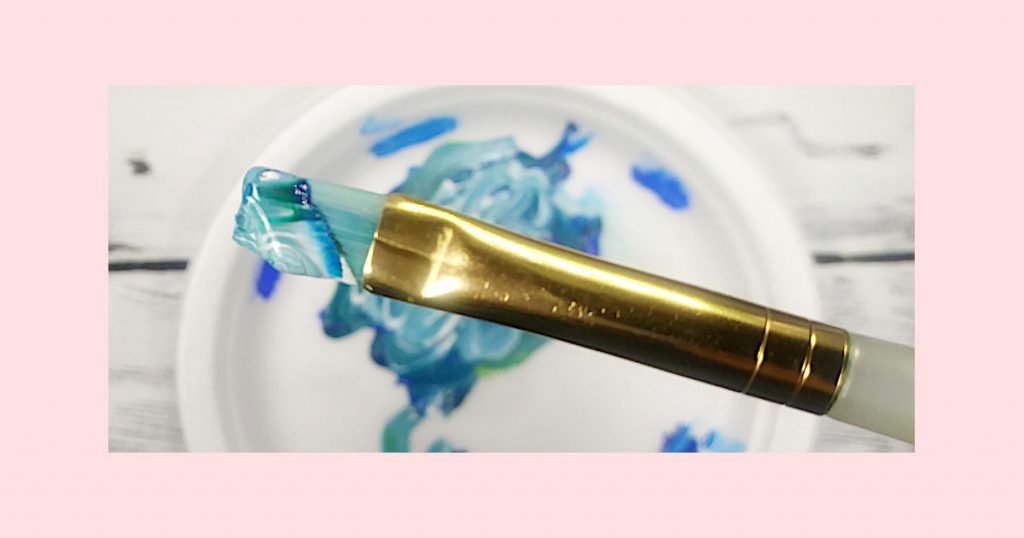 A flat paintbrush with blue, green, white, and turquoise marbled paint on it