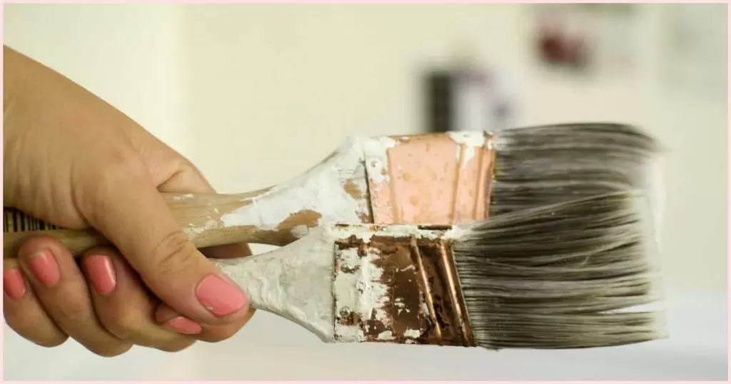 A hand with pink nail polish holding onto two large paintbrushes.