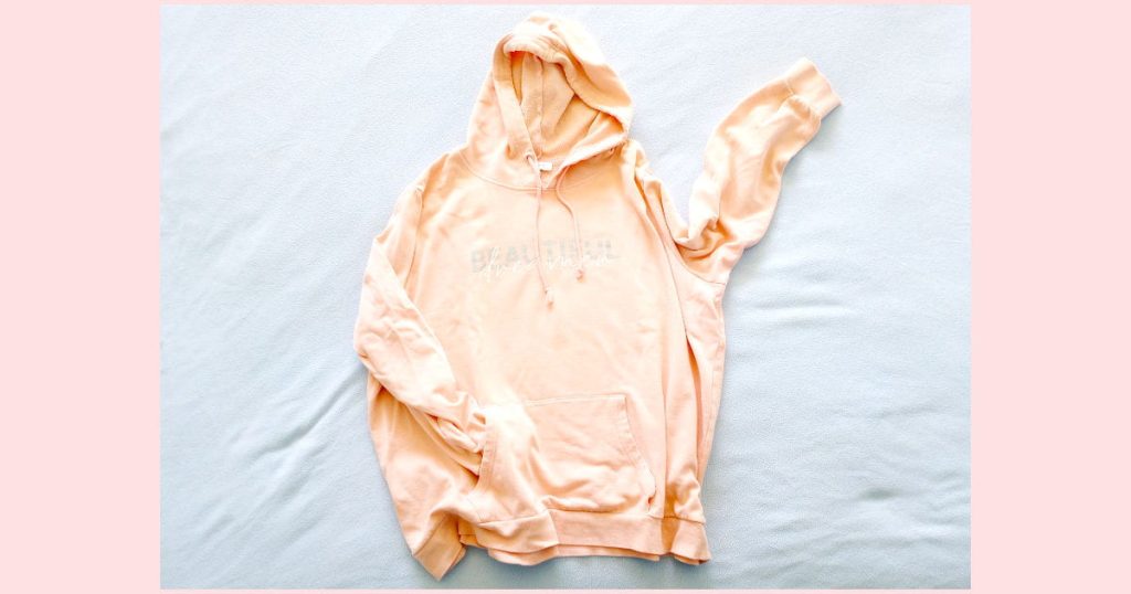 A well used peach colored hoodie that has lettering on it. It will be used to answer the question "can you use acrylic paint on fabric that has a design on it already?"