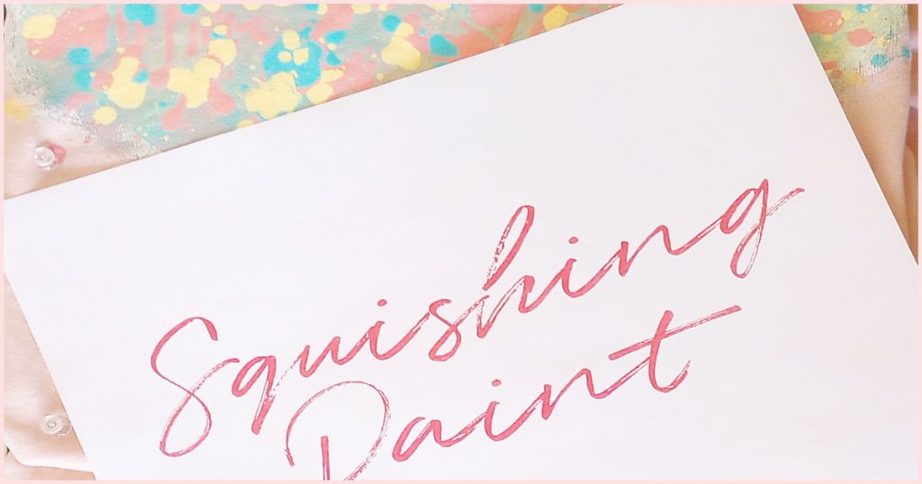 A printable of the blog name "Squishing Paint" in a red handwriting font laying on top of the hoodie being painted.
