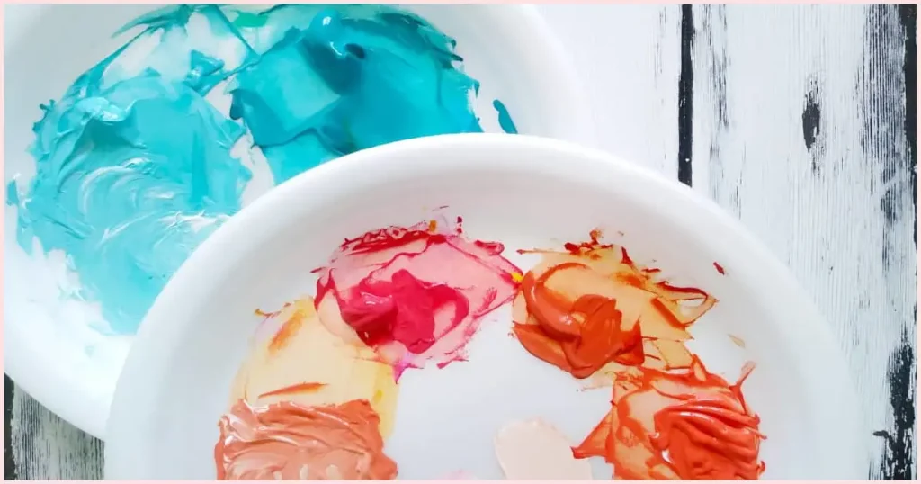 Two white round palettes, one filled with assorted turquoise paint and the other covered in various shades of pink, orange, and coral, on a whitewashed wooden slat background.