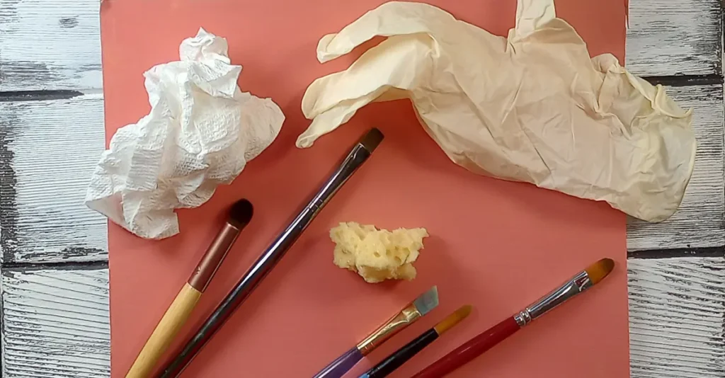Assorted brushes, a crumpled piece of paper towel, a small sponge, and a latex glove laying on top of a flamingo pink canvas. These are the tools that I'll be using to show you that painting clouds is not about the tool you use.