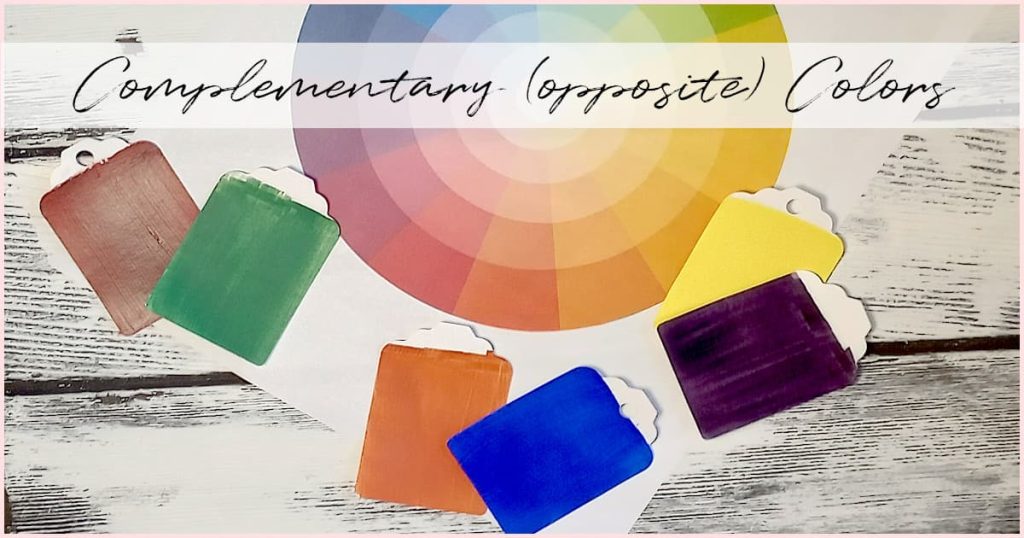 Red and green paint chips lay next to orange and blue paint chips, which lay next to yellow and purple paint chips. These are all scattered around a paper color wheel as a visual explanation of what complementary colors are on a color wheel.