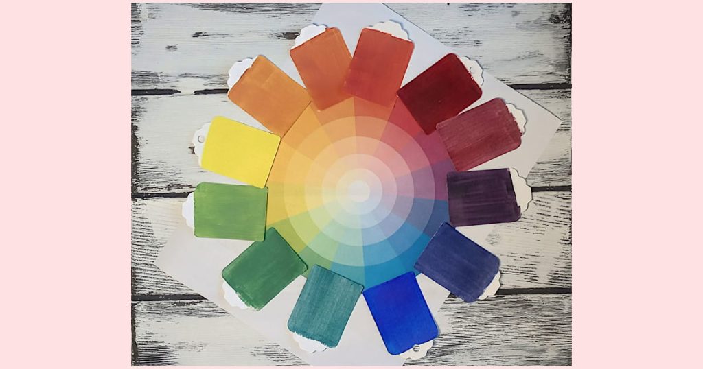 A paper color wheel with corresponding paint chips arranged around the outside of it on a white-washed wooden background.