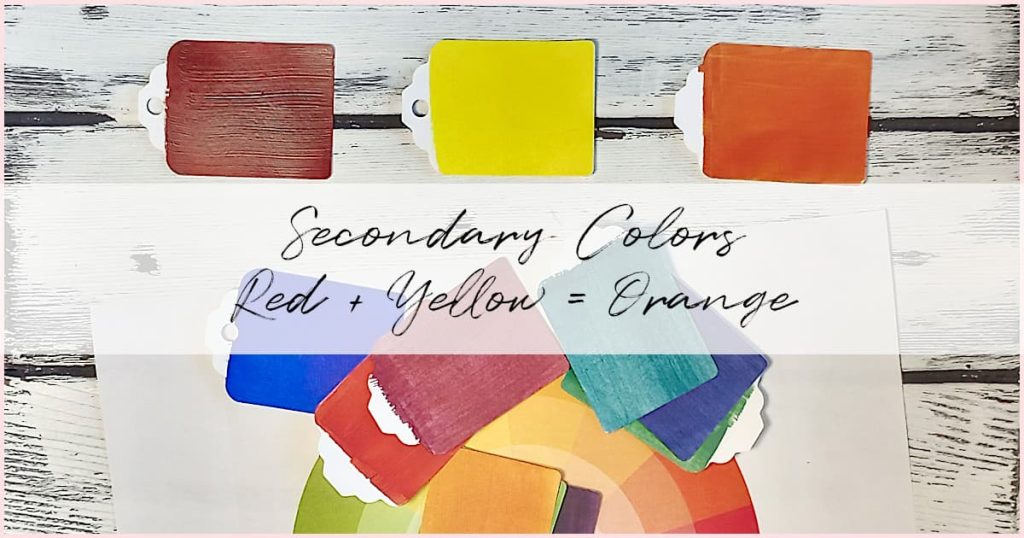 Red, yellow, and orange secondary color paint chips with an assortment of other paint chips scattered around on a white-washed wooden background.